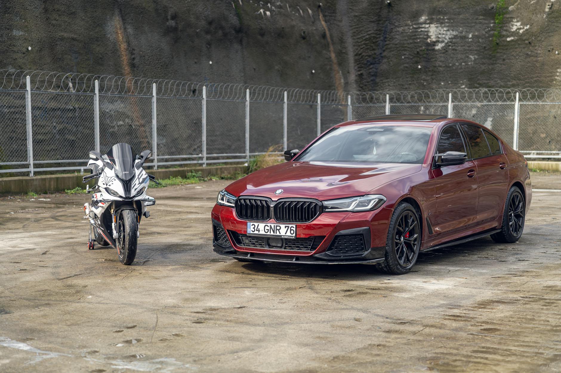 A red luxury sedan and a sports motorcycle parked on a concrete outdoor lot next to a fence.